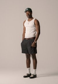 Canottiera bianca, pantaloni corti plissettati scuri, cintura intrecciata marrone, scarpe nere con lacci e inserti in mesh, calzini bianchi alti, berretto verde, sfondo neutro.
