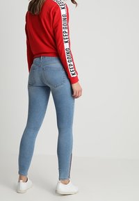 Sudadera roja con texto blanco a lo largo de las mangas, combinada con unos jeans ajustados de color azul claro y zapatillas blancas. Textura suave, diseño casual.