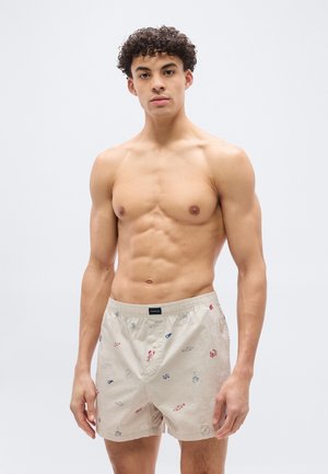 Giovane uomo con capelli ricci che indossa pantaloncini da bagno beige con stampe nautiche, in piedi contro uno sfondo chiaro e uniforme.