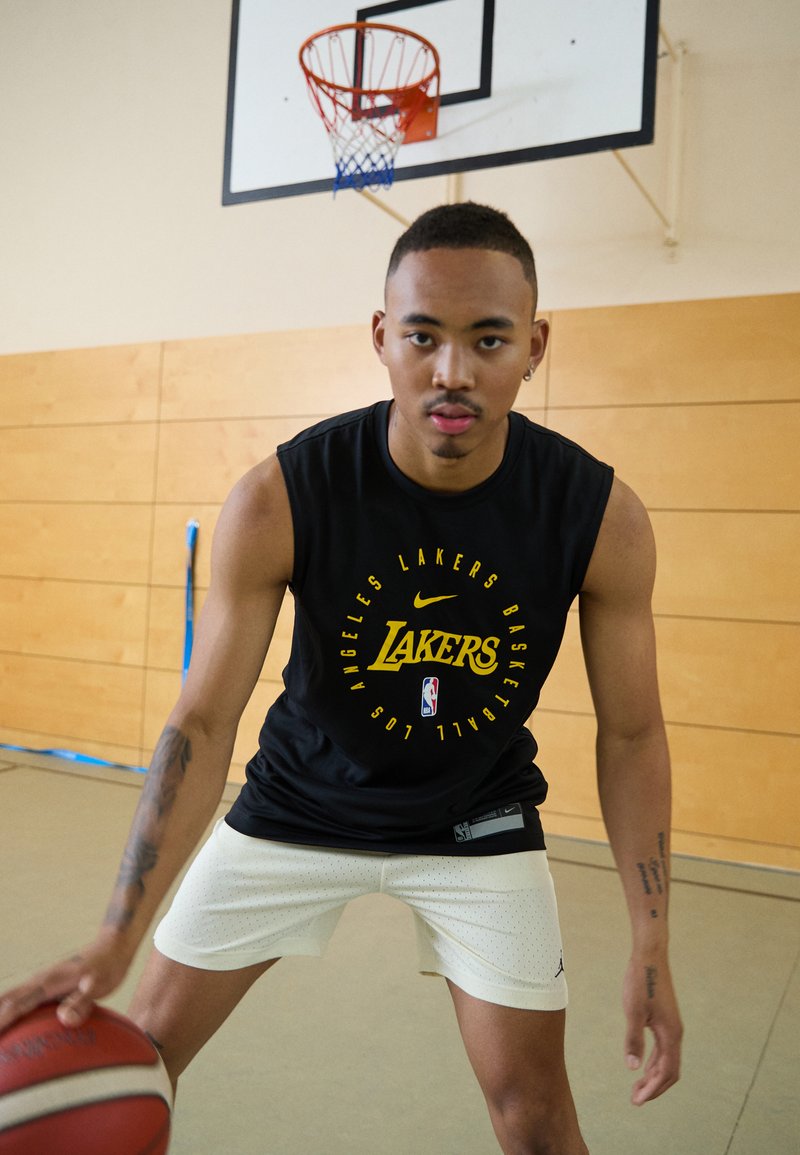 Camisola preta sem mangas com o logótipo amarelo "Lakers", calções brancos com perfurações, bola de basquete a ser driblada num ginásio.