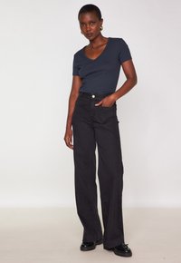 Donna con capelli corti, che indossa una maglietta navy a V, pantaloni neri a vita alta e larghi, scarpe nere e orecchini a cerchio dorati, in piedi su uno sfondo bianco.