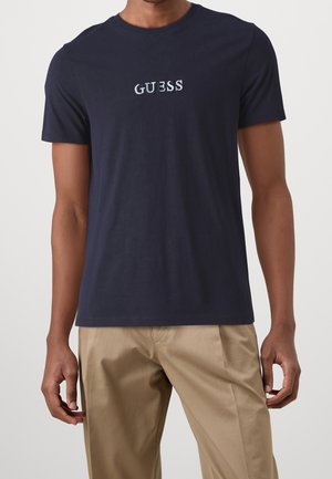 Man in een marineblauwe korte mouw T-shirt met de tekst "GUESS" op de borst en beige broek, staand tegen een effen lichte achtergrond.