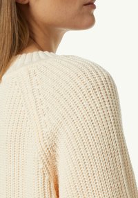Strickpullover in sanfter Cremefarbe, mit einem gerippten Ausschnitt und strukturiertem Webmuster. Der Stoff wirkt warm und gemütlich.