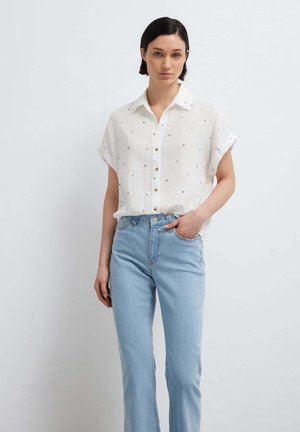 Femme aux cheveux courts et foncés portant une chemise blanche à manches courtes avec de petits pois et un jean bleu clair taille haute, main dans la poche.