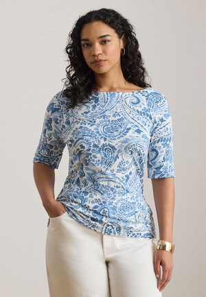 Femeie cu păr creț poartă un top albastru cu alb cu model paisley și mâneci până la cot, pantaloni albi, brățară de aur și cercei tip cerc.