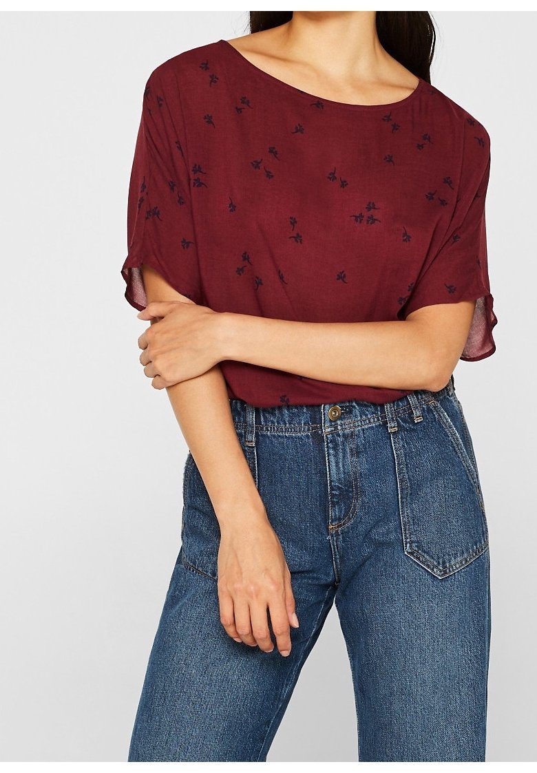 Femme portant un chemisier bordeaux à manches courtes avec de petits imprimés floraux noirs et un jean bleu taille haute, bras croisés devant.