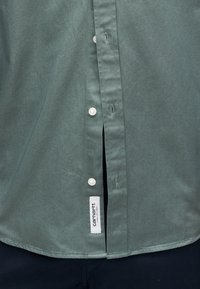 Ανοιχτοπράσινο πουκάμισο με κουμπιά, με απαλό ύφασμα, λευκά κουμπιά, πλευρικές χαραμές και ετικέτα Carhartt στο στρίφωμα.