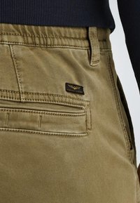 Khaki cargo kalhoty s měkkou texturou, obsahující zadní kapsy a černou nášivku s logem "American Classics" ve zlatých akcentích.