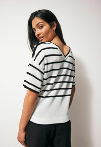 Striped knit sweater in white and black, featuring a V-neck, short sleeves, and button accents on the shoulders. Soft texture.