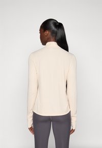 Haut beige clair à manches longues avec un col montant, texture douce et ourlet arrondi, associé à un legging gris. Détails de couture visibles aux manches.