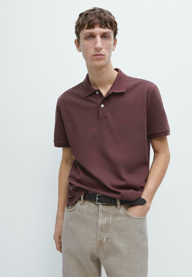 Junger Mann, der ein kurzärmeliges Polohemd in Kastanienbraun und beige Jeans mit einem schwarzen Gürtel trägt, steht vor einem weißen Hintergrund.