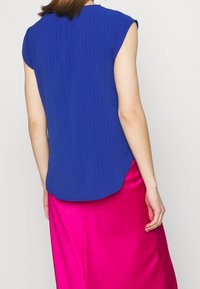 Modrý pruhovaný top s krátkými rukávy, zakřiveným lemem, spárovaný s lesklou růžovou saténovou sukní. Textura je hladká a střih je uvolněný.