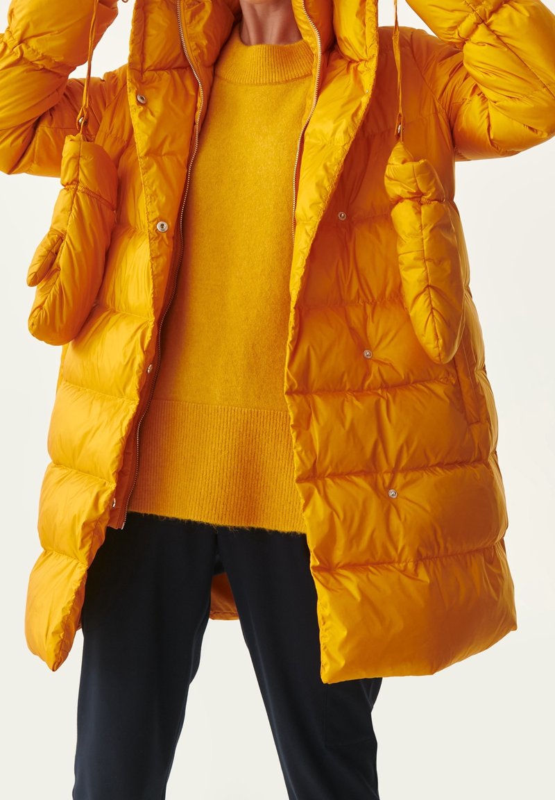 Chaqueta acolchada amarilla con capucha, que presenta una textura acolchada y cierres de botones a presión. Debajo, un suéter amarillo a juego con un dobladillo acanalado.