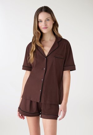 Donna che indossa una camicia marrone a maniche corte con bottoni e pantaloncini abbinati con rifiniture nere, in piedi davanti a uno sfondo chiaro e uniforme.