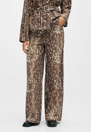 Pantalons marron à jambes larges avec un motif à pois blancs, associés à une veste ceinturée assortie, portés avec des chaussures noires et un bracelet en argent.