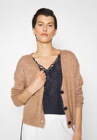 Cardigã tricotado castanho claro com mangas longas, botões frontais e punhos canelados, usado sobre uma blusa azul-marinho com detalhes em renda.