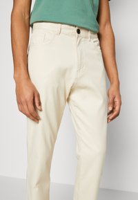 Osoba v krémových kalhotách a zeleném topu, s rukama přirozeně položenýma po stranách před jednobarevným pozadím.