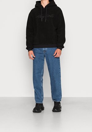 Persoon draagt een zwarte fleece-hoodie met subtiele tekst, blauwe jeans met rechte pijpen en zwarte sneakers, staand tegen een witte achtergrond.