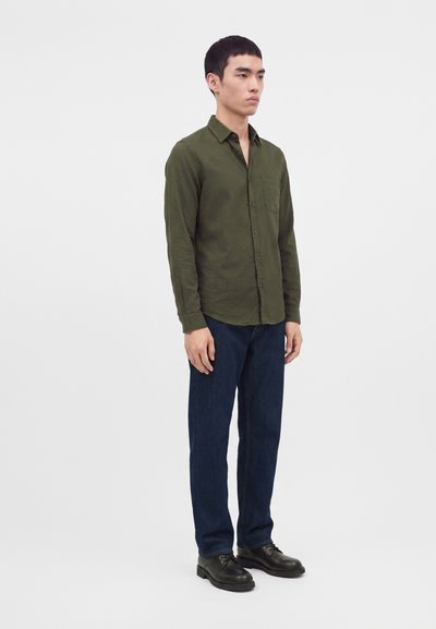 Homme debout portant une chemise kaki à manches longues boutonnée, un jean bleu foncé et des chaussures habillées noires sur un fond blanc uni.