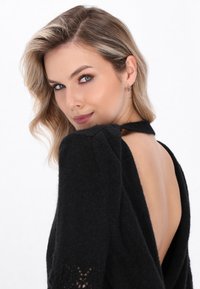 Femeie blondă cu machiaj ușor se uită peste umăr, purtând un pulover negru cu spatele deschis și cercei mici în formă de inel.