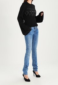 Suéter negro de punto con mangas anchas y patrones decorativos de cuentas, combinado con jeans ajustados de color azul claro y tacones de aguja negros.
