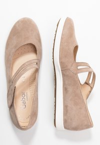 Zapatos de ante beige con punta redonda, dos correas cruzadas y detalles perforados. Presentan una suela de goma blanca y un forro interior suave.