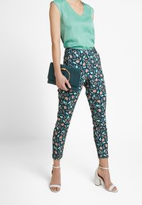 Green silk blouse, patterned trousers with blue, pink, and green botanical design, and a teal clutch with a circular handle. White block-heeled sandals.