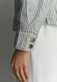 Gestreepte blauw-crèmekleurige overhemdmanchet met twee zilveren knopen. De stof heeft een gestructureerde weving, met fijne lijnen en een op maat gemaakte afwerking.