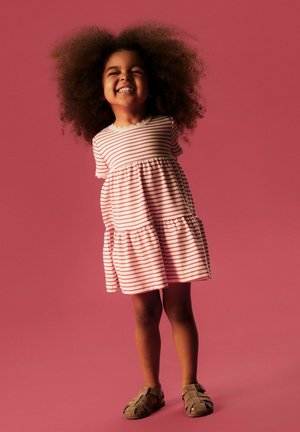 Hymyilevä nuori tyttö kiharat hiukset, raidallinen mekko ja ruskeat sandaalit, seisoo yhtenäisen vaaleanpunaisen taustan edessä.