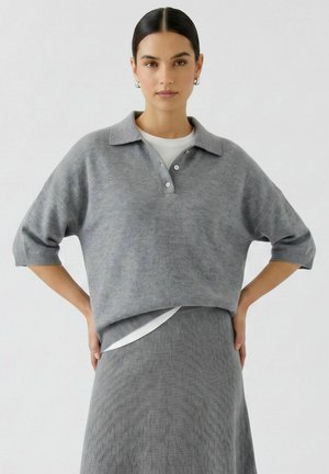 Frau mit dunklem Haar in einem niedrigen Dutt trägt einen grauen Strick-Polopullover mit Ellbogenärmeln und einem passenden Strickrock, steht mit den Händen in den Hüften.