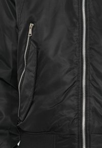 Veste bomber noire avec fermeture éclair à l'avant, poches latérales, poignets côtelés et texture de tissu lisse, mettant en avant un détail de fermeture éclair argentée brillante.