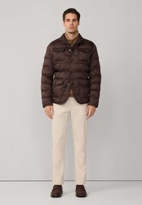 Chaqueta acolchada marrón con cierre de botones, cuatro bolsillos y cuello, usada sobre un suéter color beige, combinada con pantalones crema y zapatos marrones.