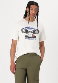 White cotton t-shirt featuring a graphic print of a woman’s face with outstretched hands, paired with olive green cargo pants.