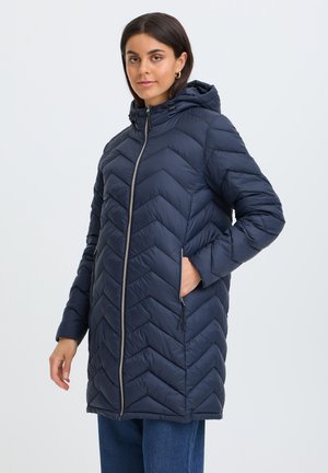 Femme portant un manteau matelassé bleu marine à capuche avec une fermeture éclair devant et des poches latérales, posant avec une main dans la poche sur un fond neutre.