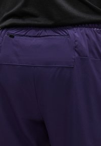 Κοντινό πλάνο σε μοβ αθλητικό σορτς με ελαστική μέση και φερμουάρ πίσω τσέπη, φορεμένο με μαύρο διάτρητο τοπ.