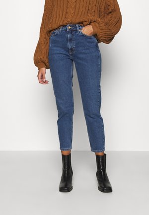 Osoba nosząca niebieskie dżinsy z wysokim stanem, brązowy sweter z grubym splotem, czarne botki, stojąca z jedną ręką w kieszeni na białym tle.