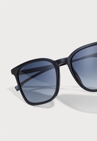 Ochelari de soare din plastic negru, cu ramă pătrată, lentile întunecate cu gradient și brandu-i minim. Au margini unghiulare și brațe subțiri.