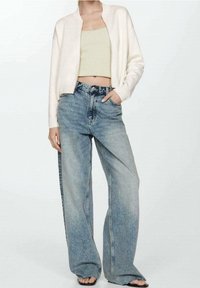 Light beige bomber jacket over a green cropped tank top, paired with loose, faded blue jeans. Model stands with one hand in pocket.