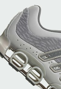 Grau sportlicher Schuh mit einem Mesh-Obermaterial, strukturierten grauen Materialien und einer klobigen weißen Sohle mit auffälligen Dämpfungselementen.