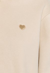 Béžový svetr vyrobený z měkké tkaniny, s malým zlatě vyšitým srdíčkem na hrudi. Jednoduchý, minimalistický styl s hladkým povrchem.