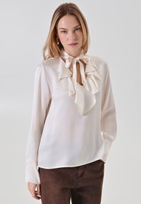Blusa crema con dettagli arricciati al colletto, tessuto morbido, maniche lunghe e un accento a fiocco annodato. Indossata con pantaloni marroni scuri.