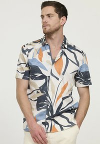 Chemise à manches courtes en tissu léger, présentant un fond crème avec des motifs de feuilles abstraits en bleu marine, orange, bleu et beige.