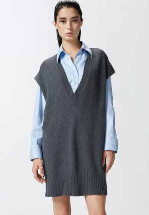 Gilet per taglie grandi in maglia grigia con scollo a V, maniche corte e orlo a costine, indossato sopra una camicia blu chiaro con colletto. Texture morbida.