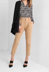 Kvinde iført tan højtaljede slim bukser, sort-hvid geometrisk mønstret bluse, sort blazer draperet over skulderen og sorte hæle.