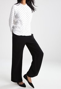 Donna che indossa una camicia bianca a maniche lunghe con pois, pantaloni neri a gamba larga e ballerine nere a punta, in piedi con una gamba incrociata.