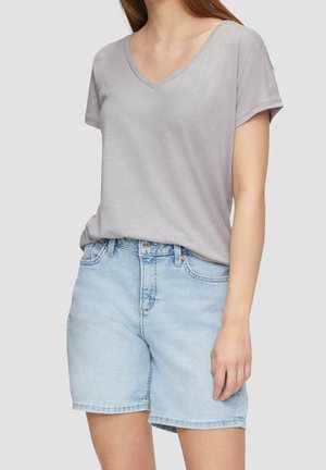 Vrouw draagt een lichtgrijs T-shirt met korte mouwen en V-hals, ingestopt in lichtblauwe denim shorts tegen een effen achtergrond.