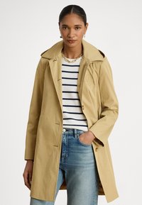 Béžový trench coat s širokým golierom, zapínaním na gombíky a dvoma prednými vreckami, vrstvený cez pruhovaný sveter a doplnený svetlomodrými džínsy.