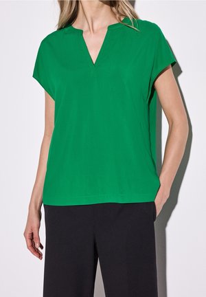 Donna che indossa una camicetta verde a maniche corte con scollo a V e pantaloni neri a gamba larga, in piedi con una mano in tasca.
