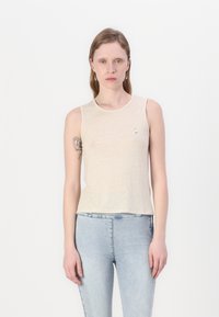 Lys beige ermeløs topp med rund hals og en liten sølvdetalj. Brukt med lyse denimjeans, som har en glatt tekstur.