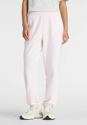 Persona che indossa pantaloni della tuta rosa chiaro e sneakers bianche, in piedi contro uno sfondo grigio uniforme.